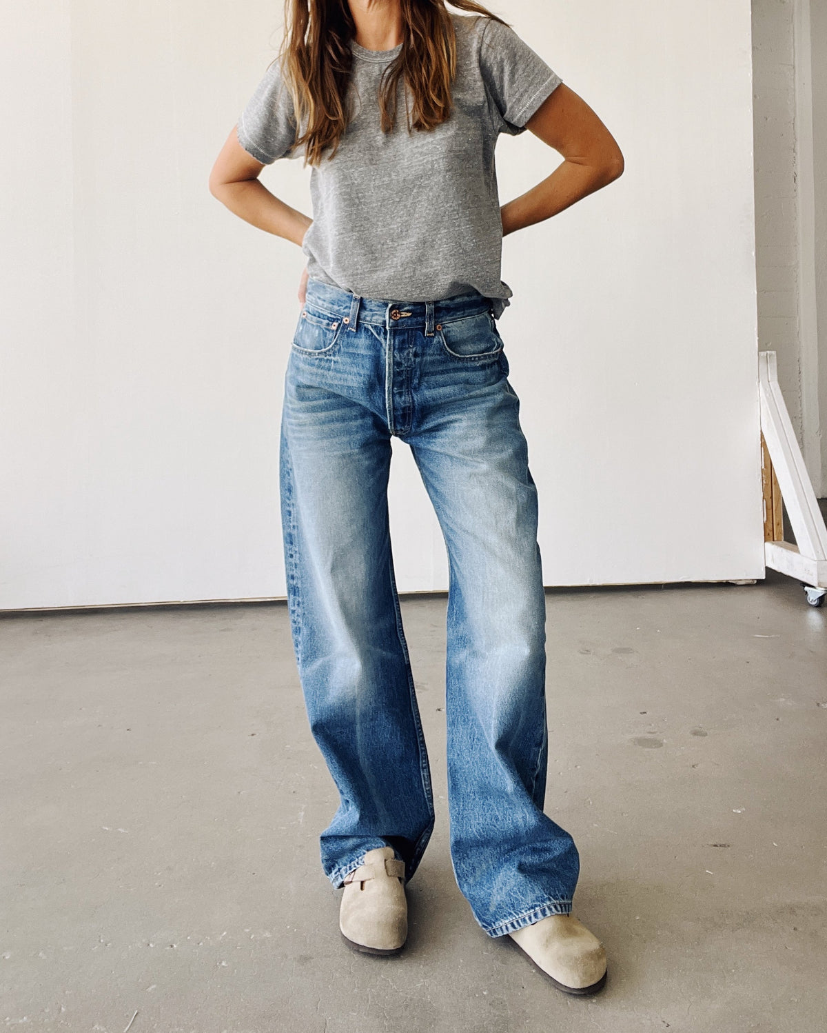 Indoors against a minimal backdrop, a person wears Hey Gang’s The Wide Slouch Jean in Harris wash, styled with a grey t-shirt and beige slip-ons. Hands on hips, their face is out of frame.