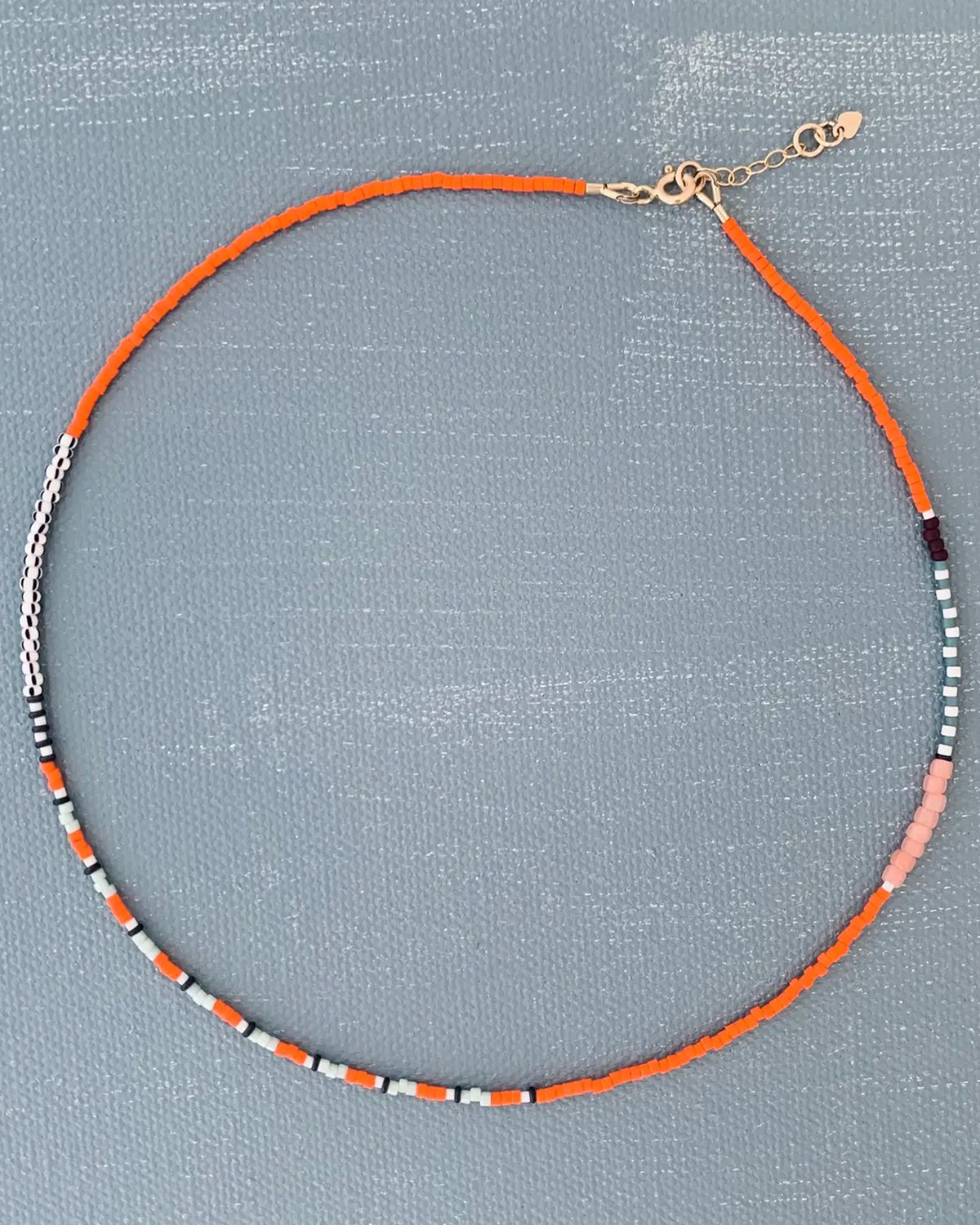 The AR Line Necklace in Marigold by Faire features Japanese glass beads in orange, white, black, and pink with a gold-filled clasp and adjustable chain, shown on a textured gray surface.