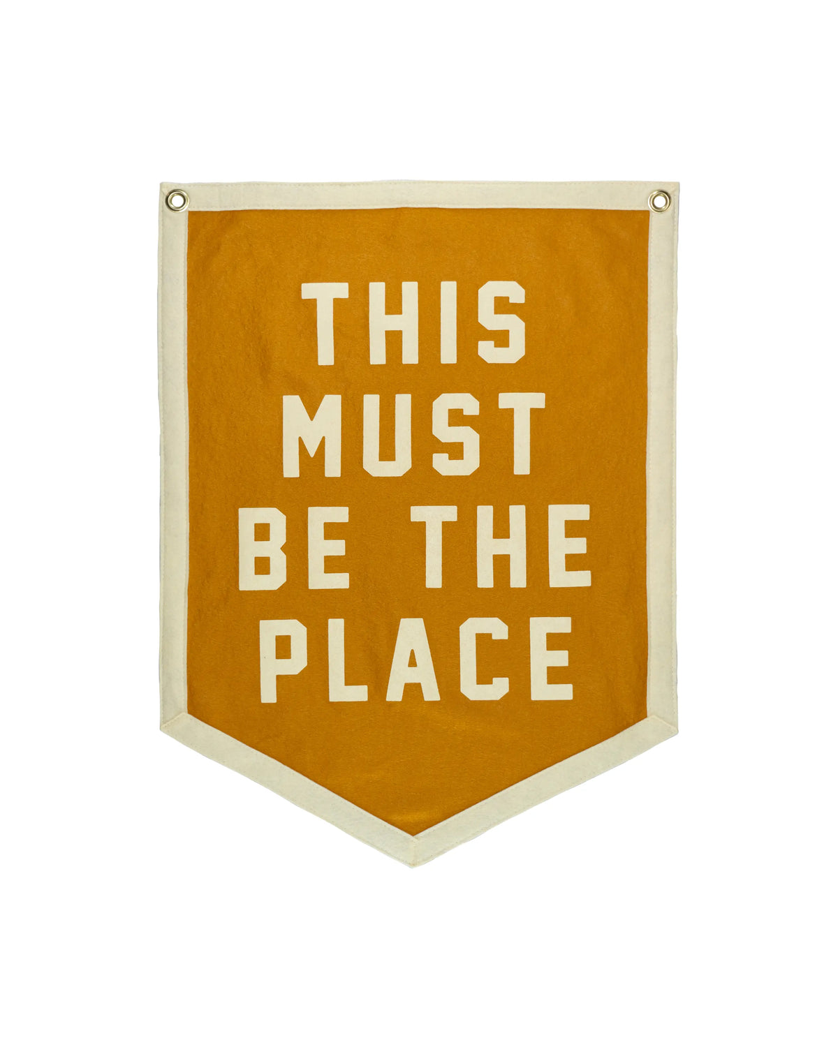 The Faire "This Must Be The Place" Camp Flag is a mustard yellow banner with white trim and bold lettering. An eye-catching gold decor piece, it features two metal grommets at the top for easy hanging.