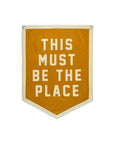The Faire "This Must Be The Place" Camp Flag is a mustard yellow banner with white trim and bold lettering. An eye-catching gold decor piece, it features two metal grommets at the top for easy hanging.