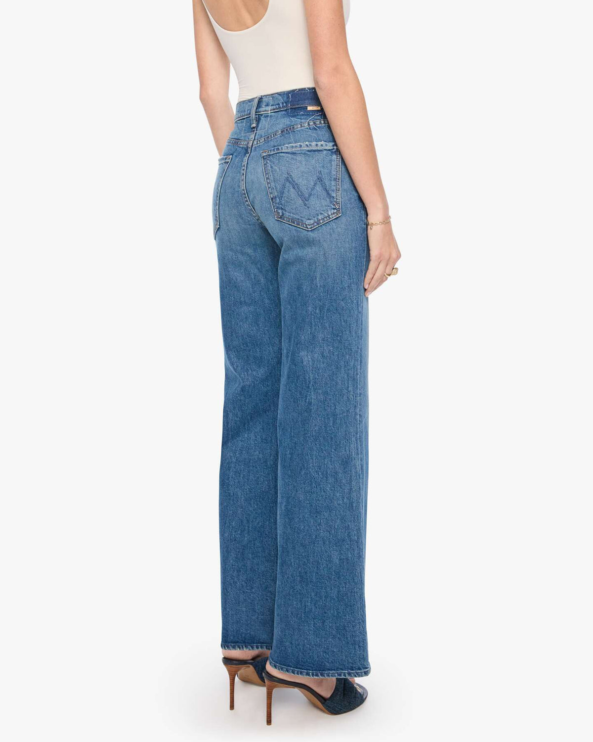 The Tomcat Roller Sneak jeans by Mother in blue denim, styled with a white tank and heels, shown from the back.