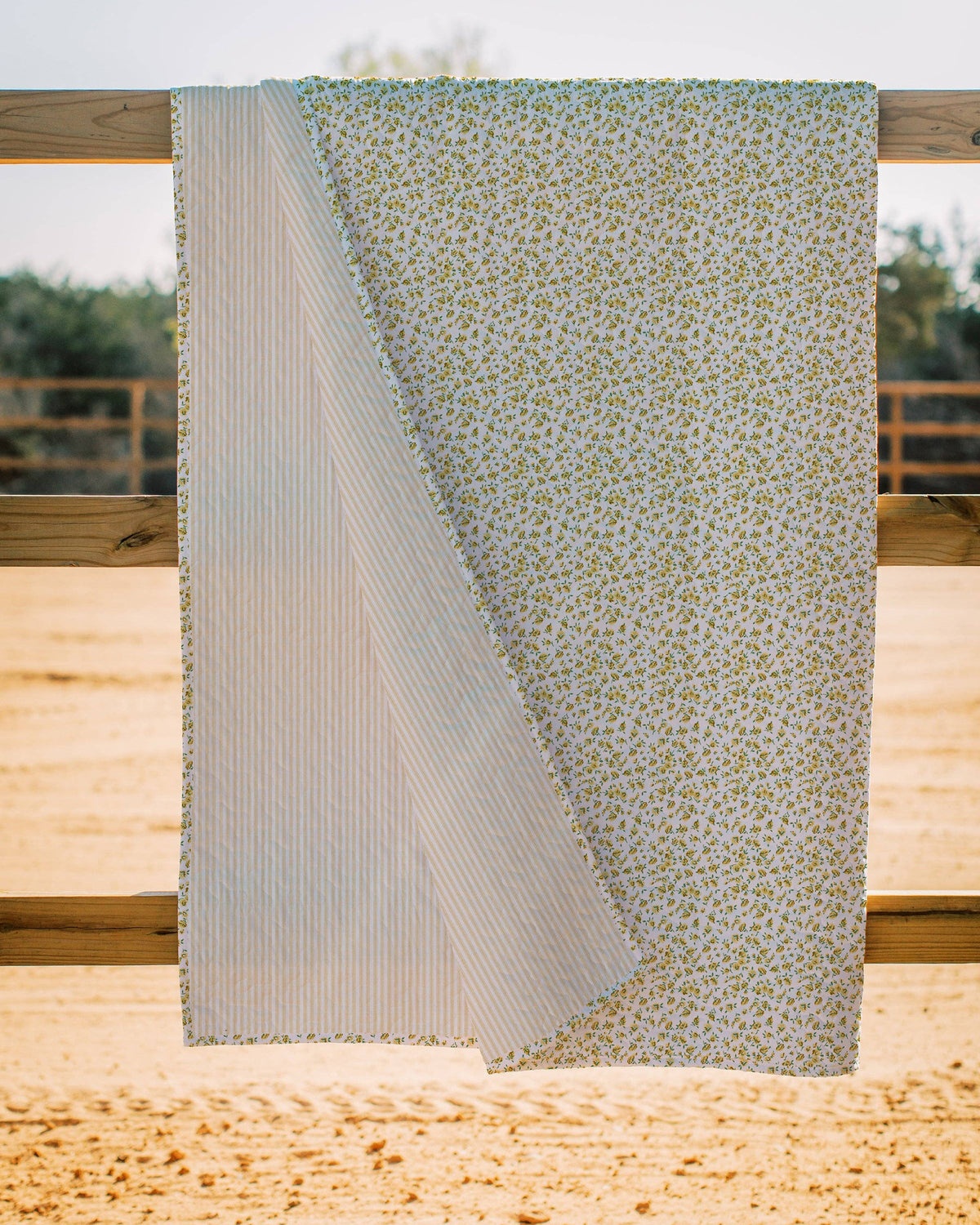 The Faire Tricia Floral Yellow Quilt, featuring a small green floral pattern on one side and thin blue and white stripes on the other, exudes cozy warmth and modern style as it hangs over a wooden fence amid sandy ground and trees.