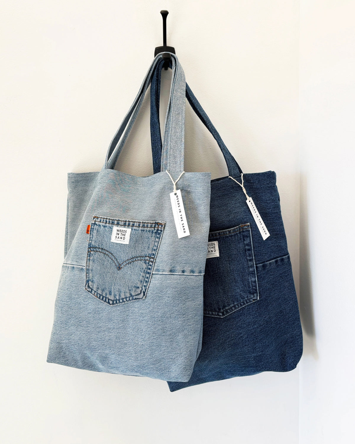 Two Faire Upcycled Denim Tote Bags, in light and dark blue, hang side by side on a black wall hook. Both have a labeled front pocket and white tag—stylish, sustainable totes ideal for everyday use.