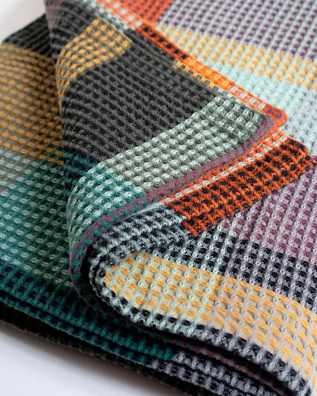 A close-up of folded Vrou-Vrou fabric by Faire highlights its bold waffle texture and colorful geometric pattern in orange, yellow, teal, black, and purple.