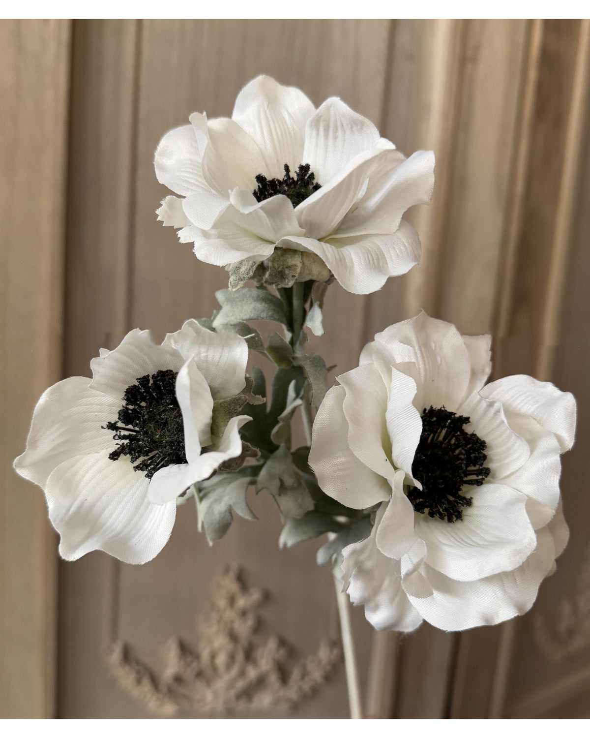 The 22'' Faux Anemone Artificial Flowers by Faire feature three silk blooms with black centers and light green stems, arranged together for a charming bouquet against a carved wooden background.