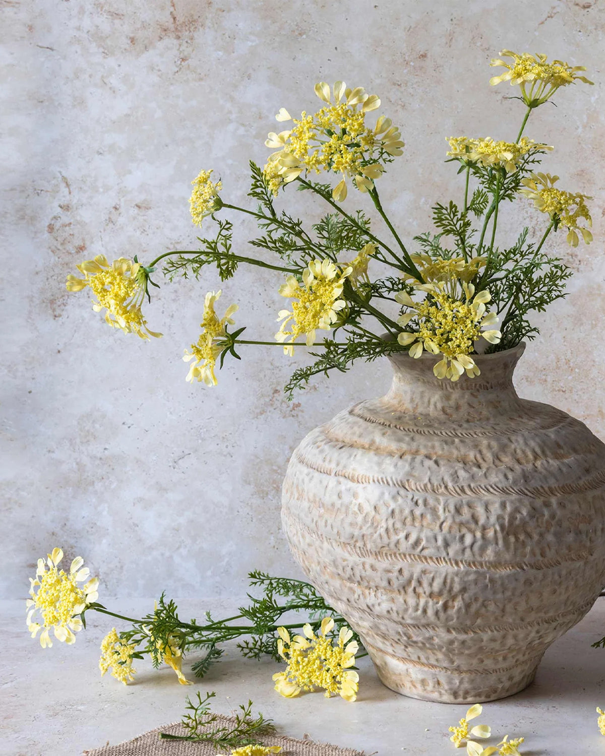 A beige ceramic vase by Abigail Ahern holds a Wild Dill faux floral arrangement with yellow and white blooms and green foliage. One artificial Wild Dill stem and small sprigs are beside the vase on a light-colored surface.