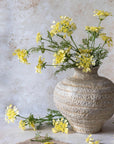A beige ceramic vase by Abigail Ahern holds a Wild Dill faux floral arrangement with yellow and white blooms and green foliage. One artificial Wild Dill stem and small sprigs are beside the vase on a light-colored surface.