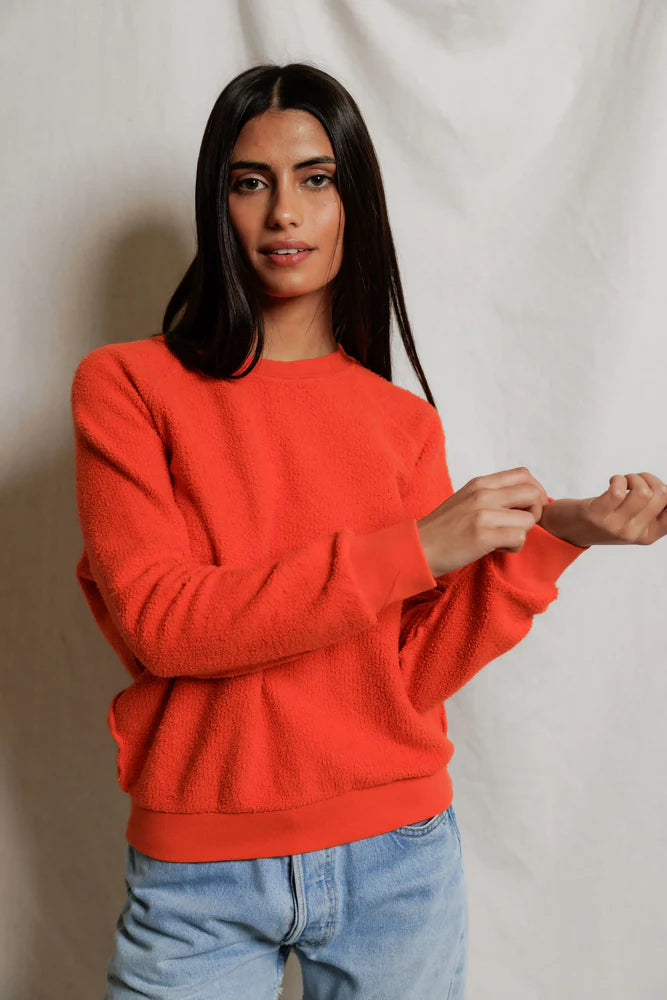 A person with long dark hair wears the Perfectwhitetee Ziggy Reverse LS Sweatshirt in bright orange cotton fleece (shrunken fit) and light blue jeans, standing against a plain light background, looking at the camera and adjusting one sleeve.