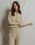 A woman with long brown hair wears the Perfectwhitetee Ziggy Reverse LS Sweatshirt in beige with matching drawstring sweatpants, standing before a light fabric backdrop and looking at the camera with a relaxed expression.