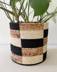 The Faire Storage Plant Basket: Zindiro features a handmade geometric checkered design in black, beige, and brown, holding a vibrant green leafy plant against a simple light background.