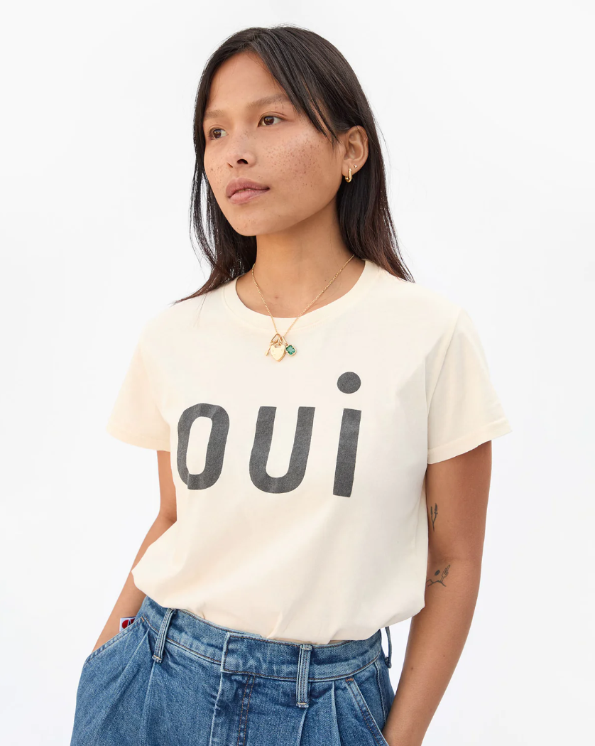 A person with long dark hair wearing a light-colored Classic Tee - OUI by Clare Vivier made in L.A. with the word "oui" written in large black letters across the front. They are also wearing blue high-waisted jeans and are accessorized with necklaces. The 100% cotton T-shirt stands out against the plain white background.