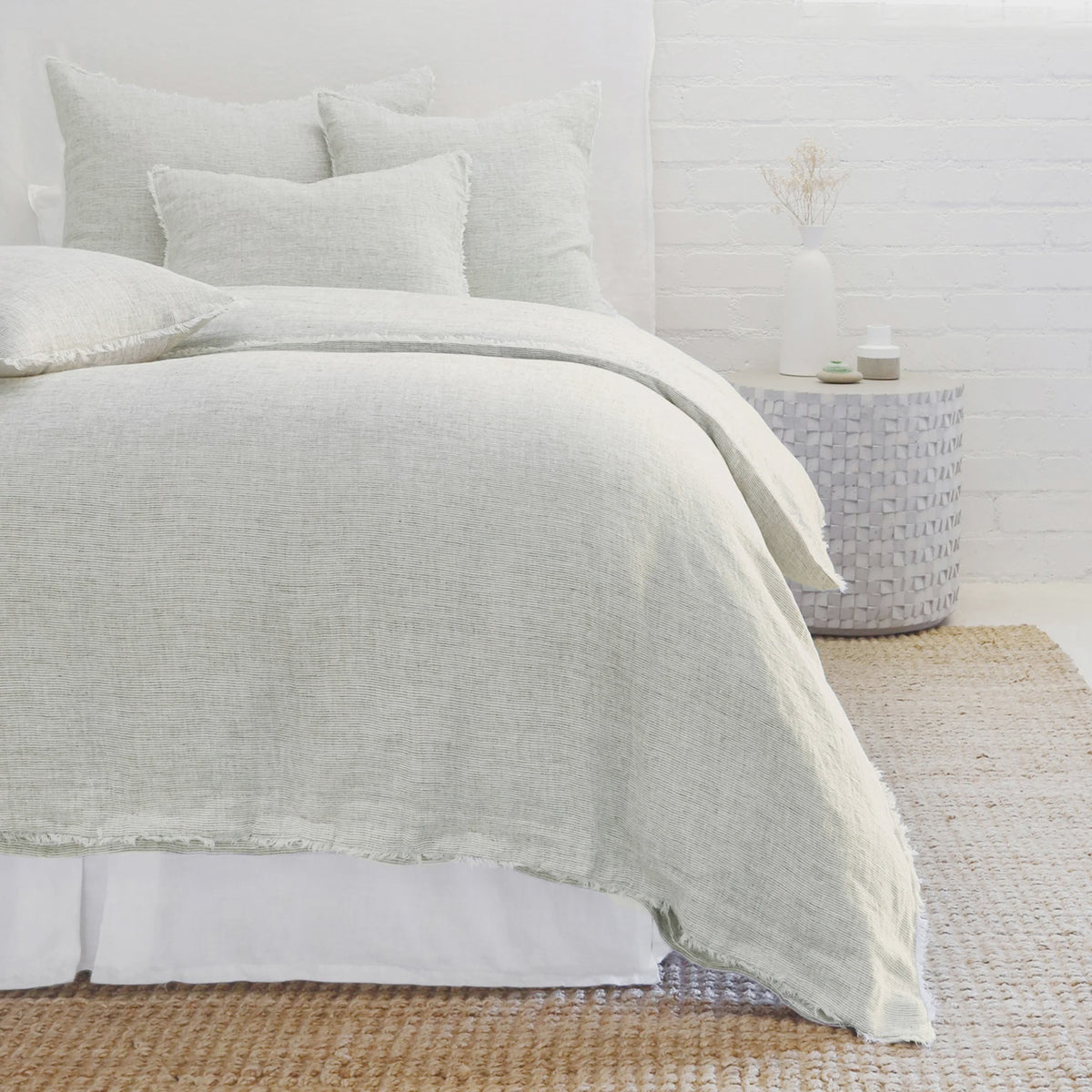 A cozy bedroom scene features a bed adorned with light gray, heathered effect bedding from the POM POM Logan Duvet collection, accompanied by matching pillows. A textured woven rug lies on the floor, and a round side table showcases subtle decor items. The room exudes a minimalist, serene ambiance.
