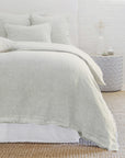 A cozy bedroom scene features a bed adorned with light gray, heathered effect bedding from the POM POM Logan Duvet collection, accompanied by matching pillows. A textured woven rug lies on the floor, and a round side table showcases subtle decor items. The room exudes a minimalist, serene ambiance.